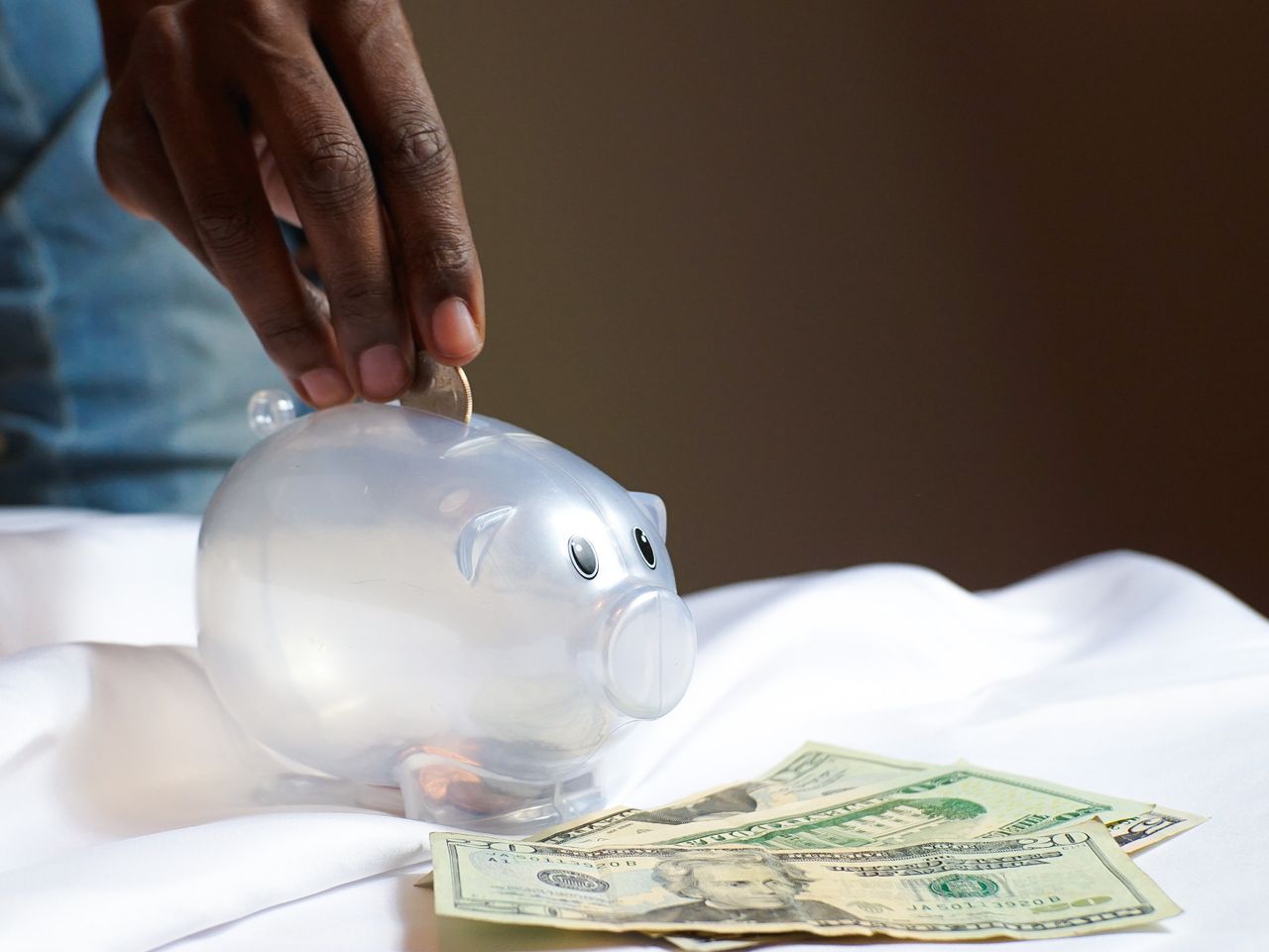 A hand placing a coin in a piggy bank with dollar bills nearby signifying savings and finance.